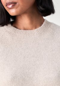 Maglione lavorato a maglia color beige chiaro con colletto rotondo, texture a costine e maniche raglan senza cuciture. Tessuto morbido e accogliente con minima variazione di colore.