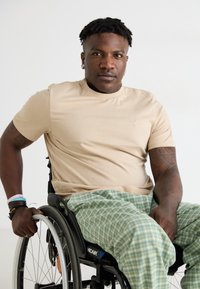 Világos bézs, rövid ujjú ing, zöld kockás pizsamanadrág, ül egy kerekesszékben, és egy tetovált karral fogja a kereket.