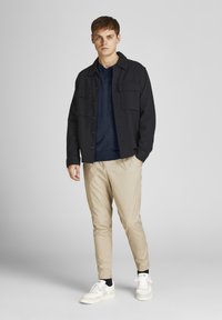 Sötét gombos overshirt, két mellzsebbel, alatta tengerészkék pulóver, krém színű, szűkített nadrág és fehér sportcipő. Egységes textúra és testreszabott illeszkedés.