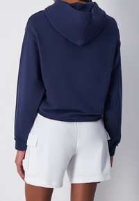 Námořnická mikina s prostornou kapucí, volným střihem a žebrovanými manžetami, doplněná o bílé šortky se bočními kapsami a texturovanou tkaninou.