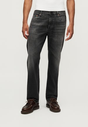 Homme portant un jean noir délavé droit et des chaussures à lacets marron foncé, se tenant devant un fond clair uni.