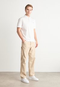 Camisa polo branca de mangas curtas, calças cargo beges com bolsos laterais e sapatilhas brancas. Design simples e casual. Textura suave.