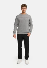 Ein Mann steht frontal und trägt einen grauen, geprägten Pullover, schwarze Hose und weiße Turnschuhe vor einem schlichten weißen Hintergrund.