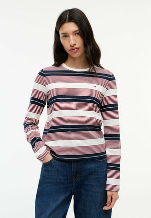 Frau mit langen dunklen Haaren, die ein weißes, marineblaues und rot gestreiftes Langarmshirt und blaue Jeans trägt, steht mit einer Hand in der Tasche.
