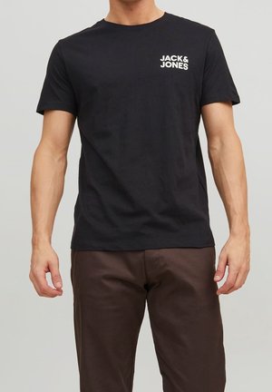 Mężczyzna w czarnej koszulce z krótkim rękawem z logo "JACK & JONES" na piersi i brązowych spodniach, stojący na neutralnym tle.