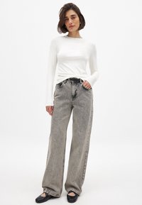 Valkoinen pitkähihainen paita yhdistettynä löysiin, haalistuneisiin mustiin farkkuihin. Farkuissa on korkea vyötärö ja suora leikkaus, jossa on näkyvät taskut.