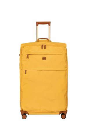 Valise souple jaune avec poignée marron, poches zippées à l'avant et quatre roulettes pivotantes, debout avec poignée télescopique étendue.