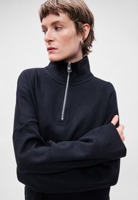 Sweatshirt preto com fecho éclair e gola alta, apresentando mangas longas e um corte descontraído. Textura suave com detalhe em zíper prateado no pescoço.