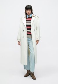 Δίπλα επάνω λευκό παλτό με μαύρα κουμπιά, φορεμένο πάνω από πολύχρωμο ριγέ πουλόβερ, ανοιχτό μπλε τζιν και καφέ μπότες.