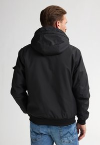 Chaqueta negra con capucha, de textura suave y puños elásticos, con cierre de cremallera y bolsillos laterales. La espalda presenta un diseño limpio y minimalista.