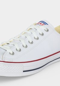 Baskets basses blanches Converse All Star avec lacets blancs, œillets métalliques, bandes rouges et bleues sur la semelle et logo sur la languette.