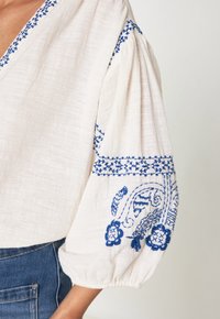 Blusa bianca con motivo ricamato blu sulle maniche e sul colletto, caratterizzata da maniche a sbuffo e un tessuto testurizzato. Materiale in cotone.