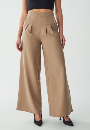 Wide-leg trousers in beige fabric with pleats, high waist, and smooth texture, paired with black pointed heels.