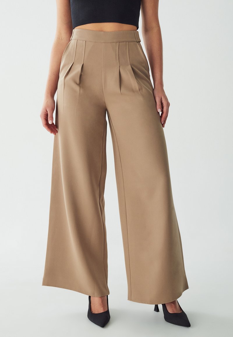 Wide-leg trousers in beige fabric with pleats, high waist, and smooth texture, paired with black pointed heels.