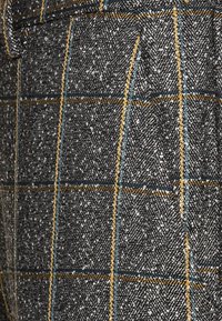 Zbliżenie na teksturowany szary materiał w wzór w jodełkę, z nałożonymi żółtymi i niebieskimi liniami w kratę, z widocznym szwem kieszeni.