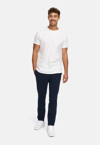 Smilende mand står med hænderne i lommerne iført en hvid kortærmet T-shirt, marineblå bukser og hvide sneakers på en hvid baggrund.