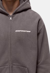 Grijze ritssluiting hoodie van zacht materiaal, met een voorzak en het "GOODFORNOTHING" logo in lichtroze over de borst.