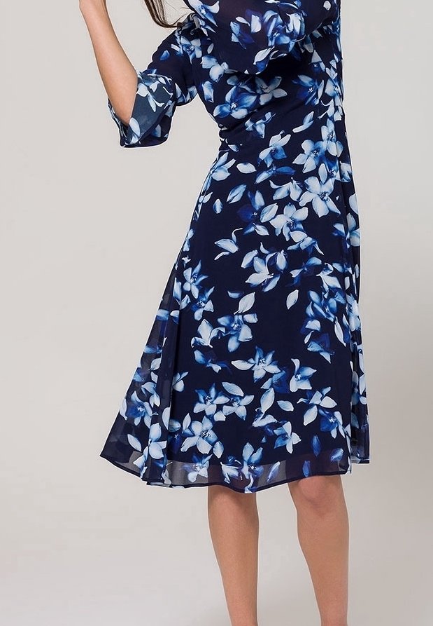Femme portant une robe marine aux motifs floraux bleus et blancs, longueur au genou, manches mi-longues, jupe évasée, debout devant un fond uni.