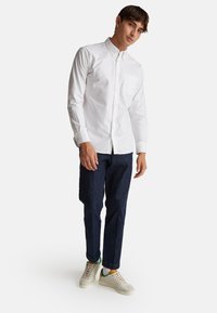 Camicia bianca a maniche lunghe con bottoni e taschino sul petto, abbinata a pantaloni slim-fit blu scuro e sneakers di colore chiaro.