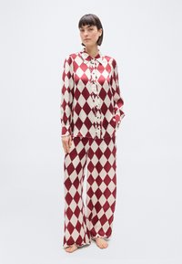 Femme portant un ensemble de pyjama en satin assorti avec un motif losange rouge et blanc, manches longues, devant boutonné, et pantalon large et ample, pieds nus.