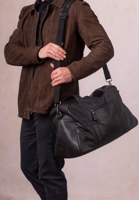 Black leather duffel bag with a zippered pocket, structured shape, and canvas adjustable strap, held by a person in a brown suede jacket.