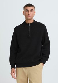 Schwarzer Strickpullover mit hohem Kragen und halbem Reißverschluss. Hat lange Ärmel und eine legere Passform. Wird über hellen Hosen getragen.
