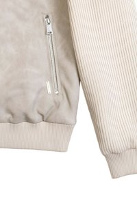 Giacca beige chiaro con un corpo liscio e textured e polsini a coste. Presenta una tasca laterale con zip e hardware metallico.