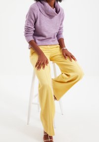 Pull col bénitier lilas en maille douce, associé à un pantalon large jaune clair, présentant une texture lisse et des détails discrets de coutures.