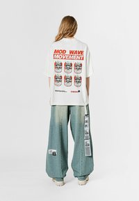 Camiseta blanca de gran tamaño con estampado gráfico que dice "MOVIMIENTO MOD WAVE", con imágenes de seis latas de refresco. Combinada con jeans azul claro y holgados.