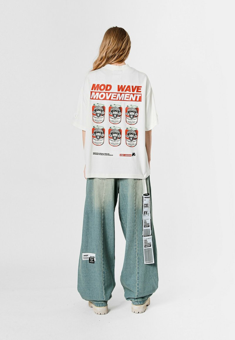 Camiseta blanca de gran tamaño con estampado gráfico que dice "MOVIMIENTO MOD WAVE", con imágenes de seis latas de refresco. Combinada con jeans azul claro y holgados.