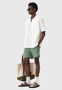 Camicetta bianca a maniche corte, pantaloni da bagno verdi con logo, sandali marroni, calzini bianchi e borsa a tracolla beige e verde, indossata da un uomo.