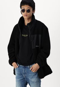 Joven con gafas de sol redondas, chaqueta de forro polar negra, camiseta gráfica negra y jeans azules, de pie con las manos en los bolsillos contra un fondo blanco.