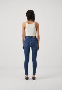 Jeans de ganga azul skinny com uma cintura alta, featuring um emblema de couro e bolsos traseiros. Combinados com uma t-shirt de tiras branca com nervuras e sapatos de salto.