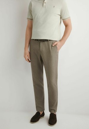 Mężczyzna ubrany w beżową koszulkę polo z krótkim rękawem, brązowe eleganckie spodnie, czarny pasek i ciemnobrązowe wsuwane buty stoi na białym tle.