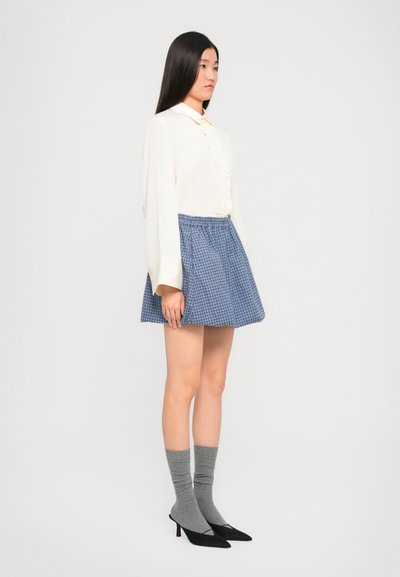 Blusa blanca con volantes, falda a cuadros azul, calcetas altas grises hasta la rodilla y tacones negros de punta. La modelo está de perfil contra un fondo liso.