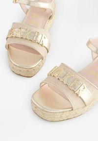 Goudkleurige platformsandalen met een geweven textuur, voorzien van een brede band met het woord "BAKER," ronde neus en espadrille-achtige details.