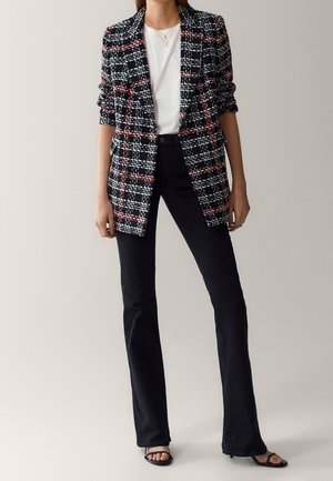 Femme portant un blazer à carreaux noir et blanc avec des accents rouges, une chemise blanche, un pantalon noir évasé et des escarpins ouverts noirs, debout devant un fond uni.