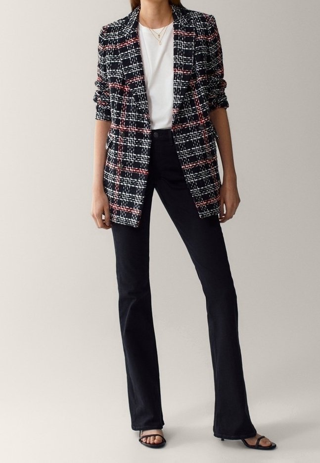 Femme portant un blazer à carreaux noir et blanc avec des accents rouges, une chemise blanche, un pantalon noir évasé et des escarpins ouverts noirs, debout devant un fond uni.