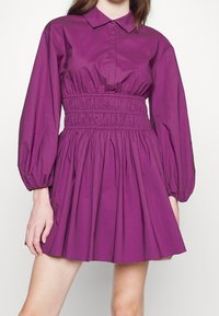 Robe violette avec un col à rabat, des manches bouffantes et une taille smockée. Jupe évasée et texture lisse avec une couleur uniforme.