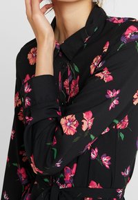 Chemise noire à motifs fleuris présentant de vibrantes fleurs roses et violettes, un col, une fermeture à boutons et un détail noué à la taille. Tissu doux avec des manches longues.