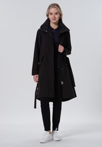 Zwarte waterdichte jas met een hoge kraag, een getailleerde taille en zijzakken. Inclusief verstelbare strik en ritsdetails. Gepaard met donkere broeken en witte sneakers.