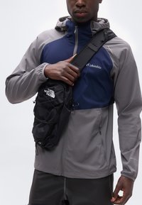 Άτομο που φοράει γκρι και ναυτικό μπουφάν Columbia και κρατά μια μαύρη τσάντα ώμου The North Face σε λευκό φόντο.