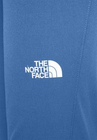 Sininen kangas, jossa on valkoinen "The North Face" -logo ja kolmasosa kaareva muotoilu, painettuna näkyvän sauman lähelle.