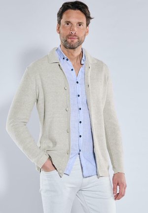 Beige gestrickte Cardigan mit Knopfleiste, getragen über einem hellblauen Hemd, kombiniert mit weißen Hosen. Der Stoff wirkt weich und texturiert.