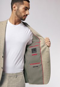 Beige Blazer mit strukturiertem Muster, ausgestattet mit roten Akzenten im Innenfutter und mehreren Innentaschen. Kombiniert mit einem weißen T-Shirt.