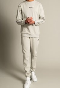 Felpa grigia chiara in cotone con logo e pantaloni della tuta abbinati, caratterizzati da polsini a coste e sneakers bianche, in piedi su uno sfondo neutro.