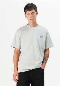 Hellgraues Baumwoll-T-Shirt mit Rundhalsausschnitt und kurzen Ärmeln. Verfügt über ein kleines blaues Textlogo im linken Brustbereich. Glatte Textur.