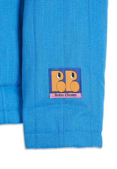 Tissu bleu avec un patch carré coloré montrant deux visages orange stylisés et le texte "Bobo Choses" en bas du patch.