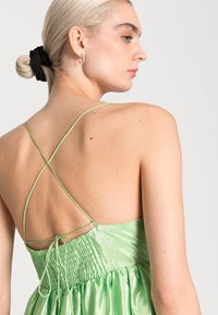 Vestido de cetim verde com corpete ajustado, costas pregueadas e alças cruzadas. O tecido tem uma textura brilhante e uma saia fluida.