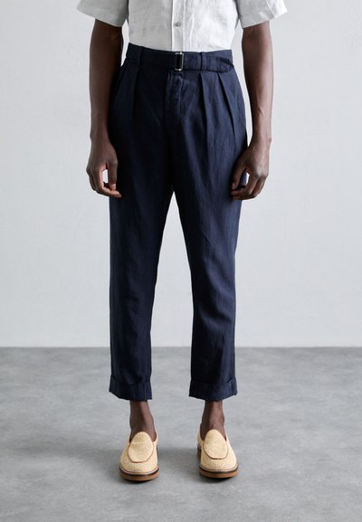 Marineblauwe, geplooide broeken met een riem en omgeslagen broekspijpen, gecombineerd met geweven instappers, op een betonnen oppervlak. Een deel van een linnen shirt is zichtbaar.